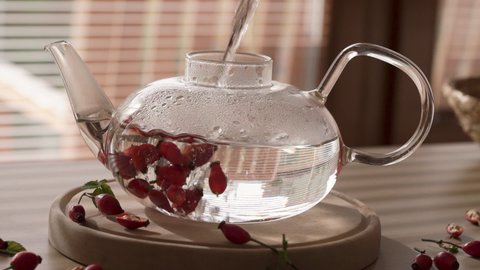 Pouring hot water into a transparent glass teapot to prepare fresh rosehip tea, slow motion
