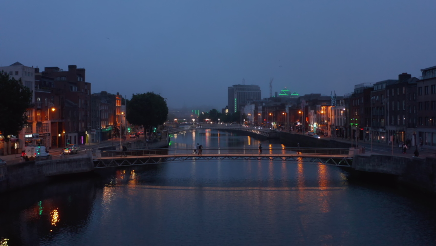 Fly over footbridges across Liffey river in city centre in evening. People enjoying night city outdoors. Dublin, Ireland
