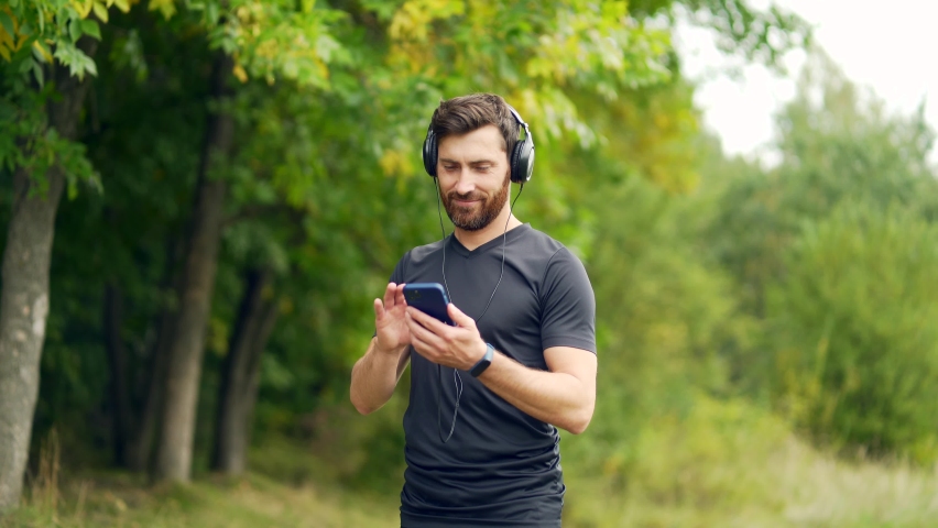young cheerful active guy sporty walking with headphones mobile phone enjoying listening to music, jogging outdoors on urban city park or forest background.  Royalty-Free Stock Footage #1081380707