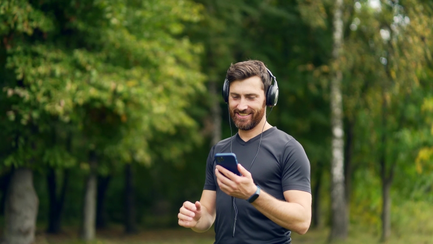 Young cheerful active guy sporty walking with headphones mobile phone enjoying listening to music, jogging outdoors on urban city park or forest background.  | Shutterstock HD Video #1081380707