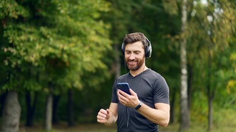 young cheerful active guy sporty walking with headphones mobile phone enjoying listening to music, jogging outdoors on urban city park or forest background. 
