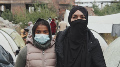 Medium close-up portrait with slowmo of Muslim woman in black veil and her 11-year-old daughter in face mask looking at camera standing at poor tent city
