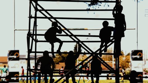Silhouette of Young Boys watching a Rugby Match from a Scaffold behind a Rugby Goal Post at Rugby Pitch. Zoom In.