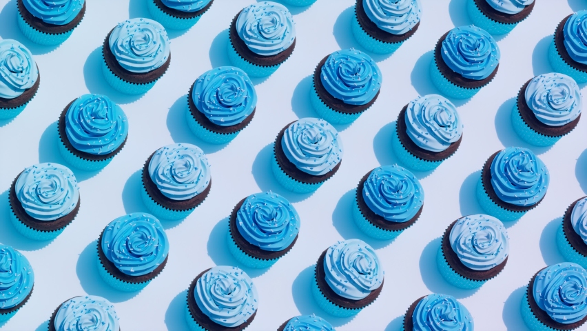 Seamless looping animation of assorted chocolate cupcakes with delicious blue cream and sweet sprinkles. Set of the homemade muffins on blue background. Decorative yummy desserts. Top view. Candy shop