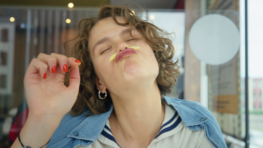 Funny and cute happy young woman giggle and smile, laugh into the camera with real and authentic emotions, play with her food, hold french potato fry under nose like moustache. Candid teenager | Shutterstock HD Video #1081385672