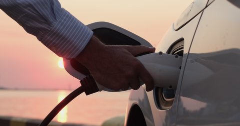A man's hand pulls out the charging plug of an electric car from the car charging socket against the backdrop of the rising sun. Dawn of the era of electric transport.