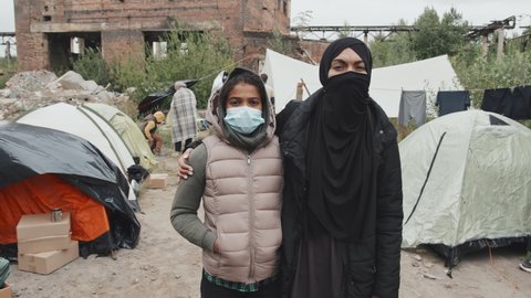 Medium slowmo portrait of Muslim woman and her 11-year-old daughter in face mask looking at camera standing at poor refugee camp where other immigrants living in tents