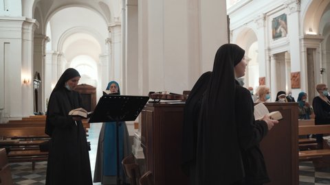 TUSCANIA, VITERBO - OCTOBER 31, 2020: Religious nuns in black cassocks holding prayer books and singing church songs during traditional mass in Tuscania village, province Viterbo. Religious sisters