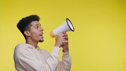 Excited young African American man 20s looking aside screaming in megaphone. Guy in casual light sweat shirt posing isolated on yellow background studio. People lifestyle concept. Copy space mock-up