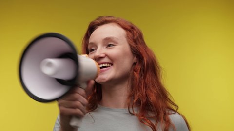 Smiling Young woman screaming in megaphone wearing basic green shirt posing isolated on yellow background studio. Ginger hair Caucasian girl screaming in megaphone. Zoom out