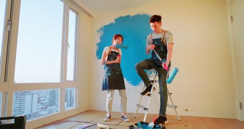 young asian couple decorate home - they painting wall with blue color together and singing with dance for fun