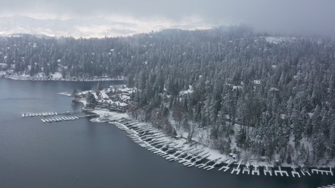 Winter season snowy mountain forest aerial shot. Breathtaking natural landscape, frozen forest and dark mountain lake. Wonderland village with boat landing docks at waterfront forest cabins, Arrowhead