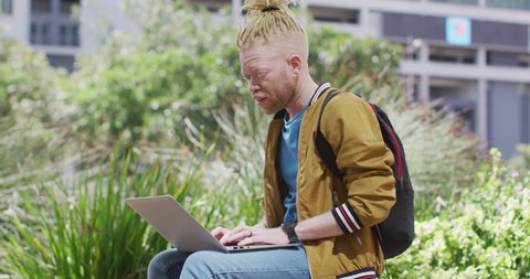 Thoughtful albino african american man with dreadlocks sitting in park using laptop. digital nomad on the go, out and about in the city.