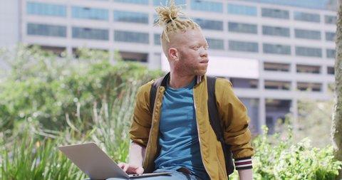 Thoughtful albino african american man with dreadlocks sitting in park using laptop. digital nomad on the go, out and about in the city.