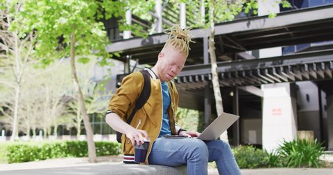 Thoughtful albino african american man with dreadlocks sitting in park drinking coffee, using laptop. digital nomad on the go, out and about in the city.
