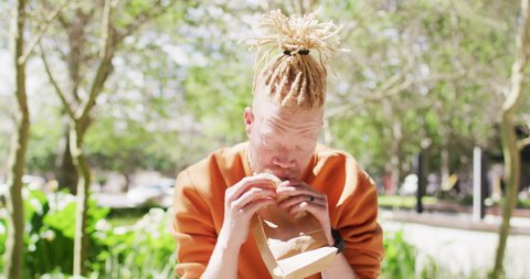 Albino african american man with dreadlocks sitting in park with bike eating sandwich. on the go, out and about in the city.