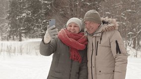 Medium of happy senior Asian woman and Caucasian man wearing winter clothing, video calling using smartphone, standing in snowy park