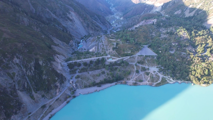 View of the mountain lake Issyk from a height. Turquoise-emerald water from the glacier. Reflection of mountains, forests and the sun on the water. The river flows into the lake. Issyk Dam. Kazakhstan