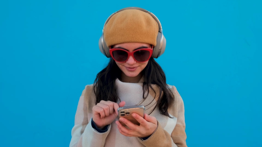 Positive female with black hair in sunglasses and hat listening to music in headphones while surfing cellphone on blue background