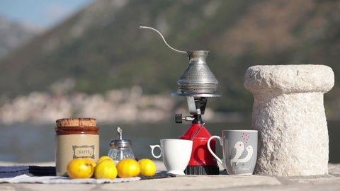 Red camp gas burner and coffee cezve at picnic on old pier in bay. Coffee crockery still life near ancient stone bollard on pier with mountain blurred background. Donji Stoliv, Montenegro, 10 28 2021