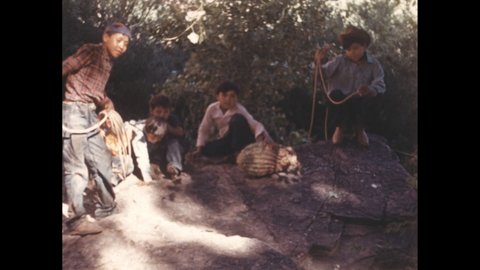 1940s: Boys slinging lassos at rolling gourd. Children playing on rock.