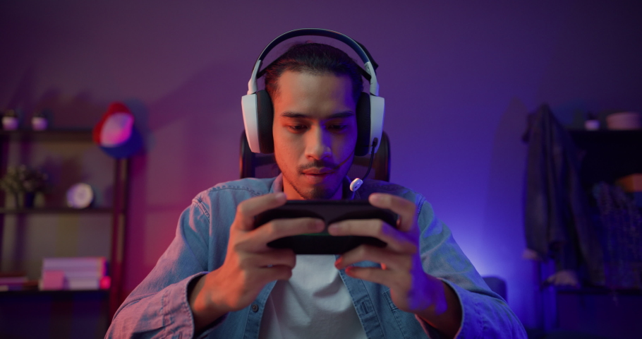 Happy asia man gamer wear headphone competition play video game online with smartphone colorful neon light in living room at night modern house. Esport streaming game online, Home quarantine activity. | Shutterstock HD Video #1081417127