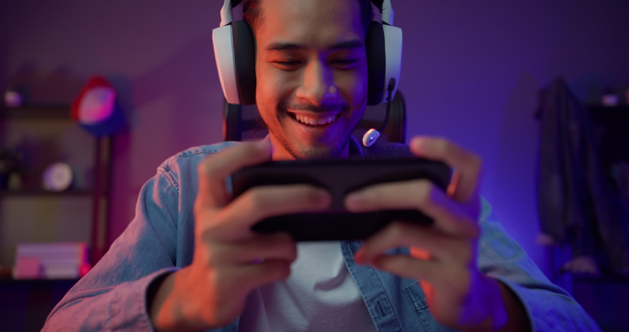 Happy asia man gamer wear headphone competition play video game online with smartphone colorful neon light in living room at night modern house. Esport streaming game online, Home quarantine activity. | Shutterstock HD Video #1081417127