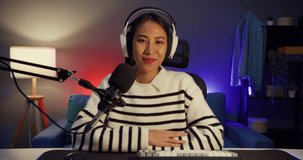 Happy asia lady blogger music influencer looking at camera broadcast record wear headphone online live talk in microphone with audience in living room house studio at night. Content creator concept.