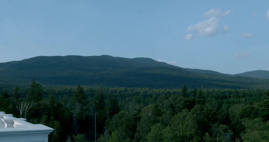 A long panning shot from the roof of a hotel overlooking the white mountains of New Hampshire. The mountains loom over a forest and golf course under a blue sky.