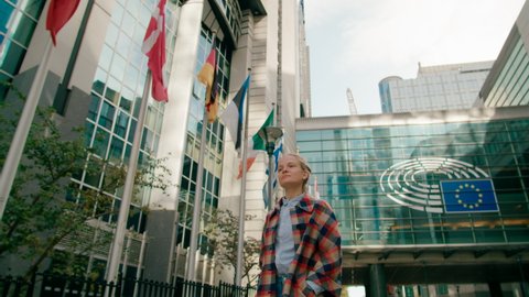 Woman Walks along European Flags near EU Parliament Modern Office Building in Brussels, Belgium. Politics, Economy and Business Concept. 4K gimbal follow low angle shot in slow motion