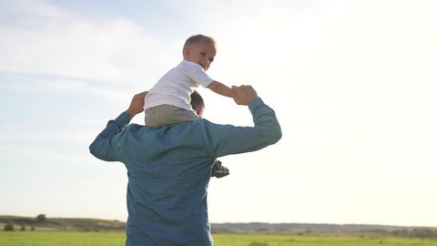 happy father walks with little son in park in summer, holds him on his shoulders. cute toddler enjoying his father's back ride outdoors. dad rolls happy toddler, son on back on street.
