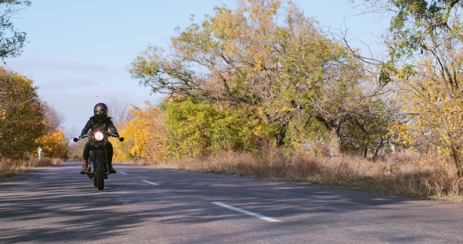 Woman biker with long blonde hair in black helmet rides motorcycle at high speed along road between colorful trees in autumn slow motion