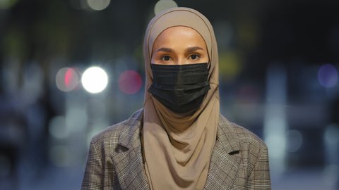 Close-up islamic female in medical mask posing in night city. Muslim business woman wearing hijab masked girl standing looking at camera evening urban background outdoors pandemic coronavirus covid