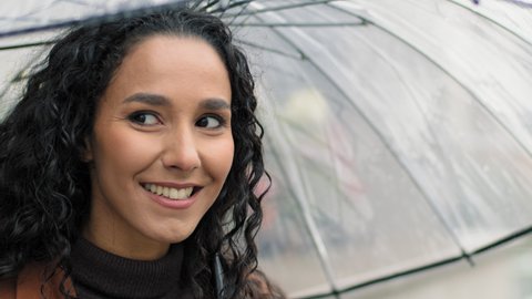 Portrait carefree attractive beautiful woman female happy hispanic girl with long dark curly hair holding transparent umbrella standing in city in rainy weather rain smiling toothy looking at camera