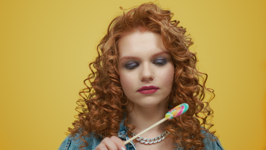 Closeup attractive woman holding colorful lollipop in studio. Portrait of smiling girl covering eye with sweet candy stick. Redhead lady eating sugar dessert food having fun flirt on yellow background