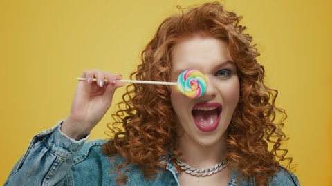 Closeup attractive woman holding colorful lollipop in studio. Portrait of smiling girl covering eye with sweet candy stick. Redhead lady eating sugar dessert food having fun flirt on yellow background