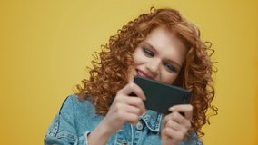 Closeup excited woman playing game online smartphone at yellow background. Redhead girl using phone. Portrait of happy female winner winning video game on cellphone. Lady celebrating victory success