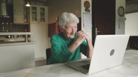Slow motion - Old grannie having a video call with loved one through her laptop computer and laughing out loud. Modern senior female socializing through the web