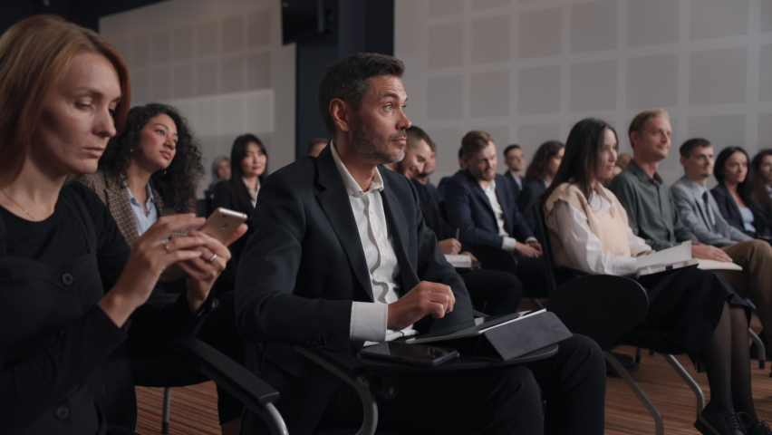 Group of people listens speech at business conference. Stage talk at summit meeting in crowded modern hall. Caucasian man training at economy forum. Business work of sitting male person in suit indoor