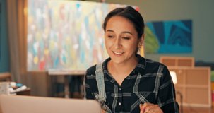 Young beautiful artist sits in studio in front of laptop screen, talks via webcam, social network with friend, describes her new painting, painting vision, shows found paints