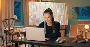 Artist, dressed in work clothes, sitting in studio, behind painting on canvas standing on easel, girl has laptop in front of her, she is talking through social media via webcam with manager