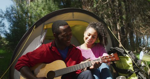 Smiling diverse couple sitting in tent and playing guitar in countryside. healthy, active lifestyle and outdoor leisure time.
