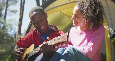 Smiling diverse couple sitting in tent and playing guitar in countryside. healthy, active lifestyle and outdoor leisure time.