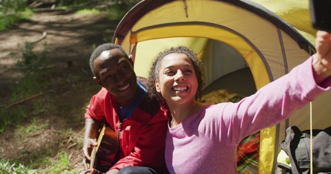 Smiling diverse couple sitting in tent and taking selfie in countryside. healthy, active lifestyle and outdoor leisure time.