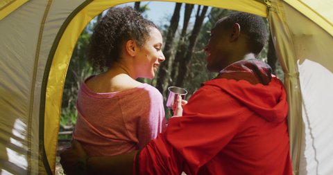 Smiling diverse couple sitting in tent and drinking tea in countryside. healthy, active lifestyle and outdoor leisure time.
