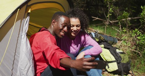 Smiling diverse couple sitting in tent and taking selfie in countryside. healthy, active lifestyle and outdoor leisure time.