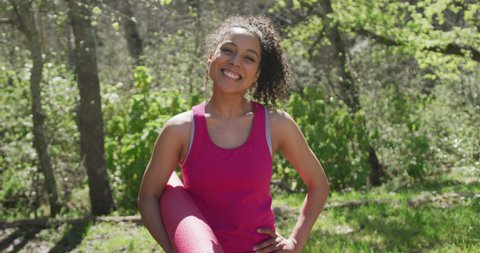 Portrait of smiling biracial woman wearing sportswear in forest with yoga mat. healthy, active lifestyle and outdoor leisure time.