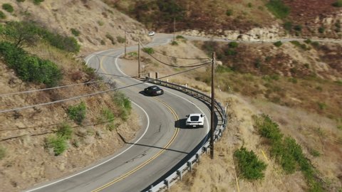 Fast speedy cars racing in Los Angeles suburban, California USA 4K aerial view. Black and white exotic sports car driving by the winding road in Malibu canyon high above Pacific ocean on background