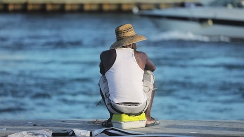 Waterfront Miami fishing harbor June 2019. Athletic African American guy fishing at sunset. Rear view of man in white T-shirt and straw hat at water background. Slow motion yacht sailing at sunset Editorial Stock Video