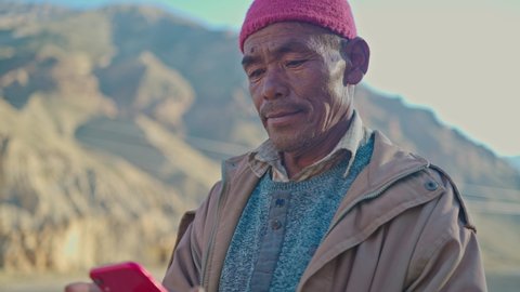 Close shot of an Asian old man wearing a head cap and winter jacket is standing and using a smartphone to type a text message in the cold barren mountainous region